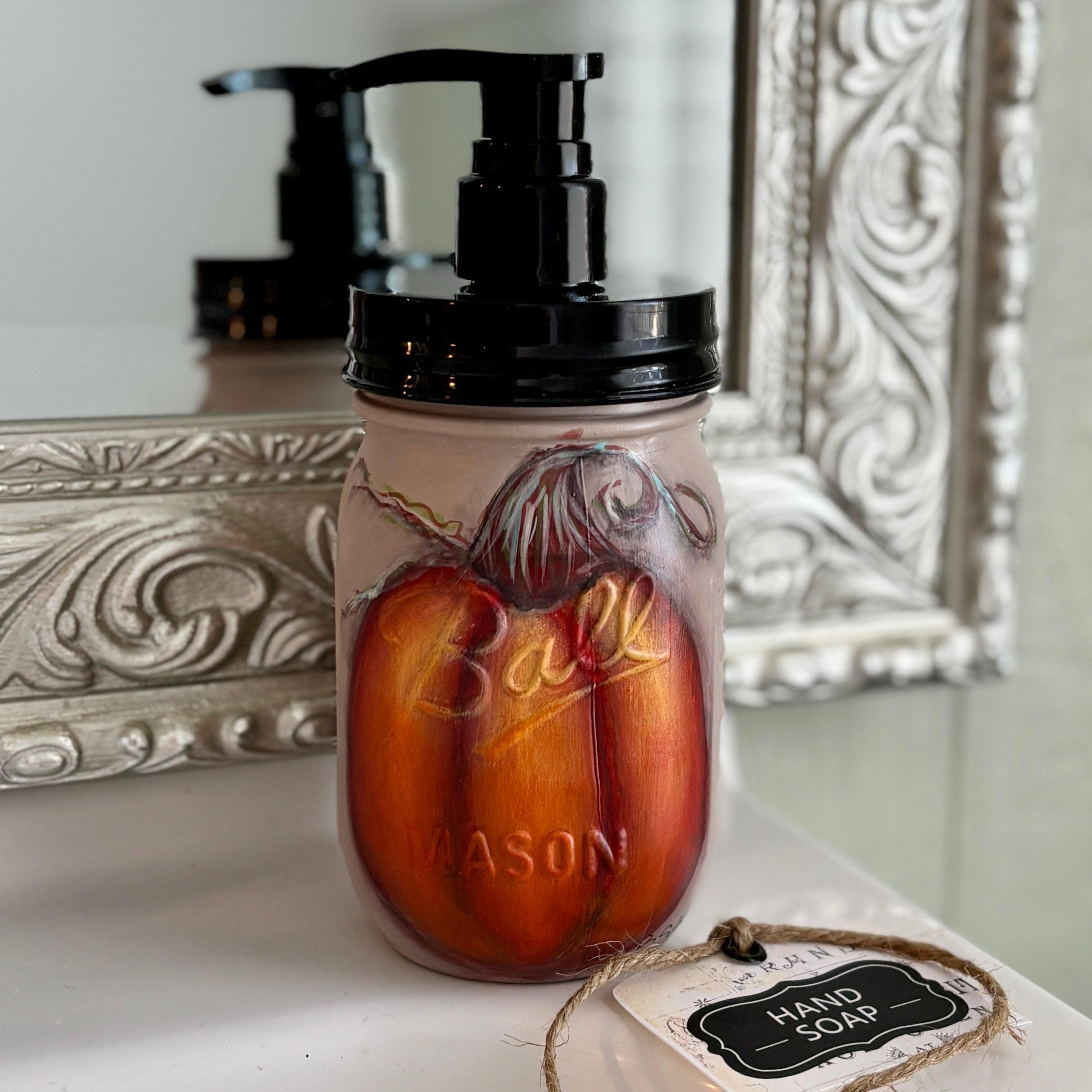 A decorative hand soap dispenser styled like a mason jar with a black pump, featuring a painted orange and red pumpkin design on a light background. A small paper tag reading 'HAND SOAP' is tied to the front, and an ornate silver mirror frame is visible in the background.